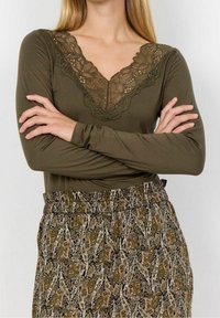 Olivenfarbener Langarm-Top mit einem Spitzenausschnitt in V-Form, kombiniert mit gemusterten, elastischen Hosen mit aufwendigen Paisleymotiven.