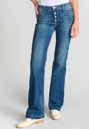 Flared blue denim jeans with a five-button front closure, subtle whiskering, and stitched detailing on pockets. Paired with white loafers.