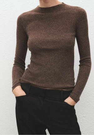 Personne portant un haut marron ajusté à manches longues côtelé, rentré dans un pantalon noir taille haute, les mains dans les poches.