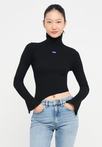 Maglione a collo alto nero a costine con maniche lunghe e design cropped. Con una patch blu del logo Hugo sul petto. Indossato con jeans a vita alta.