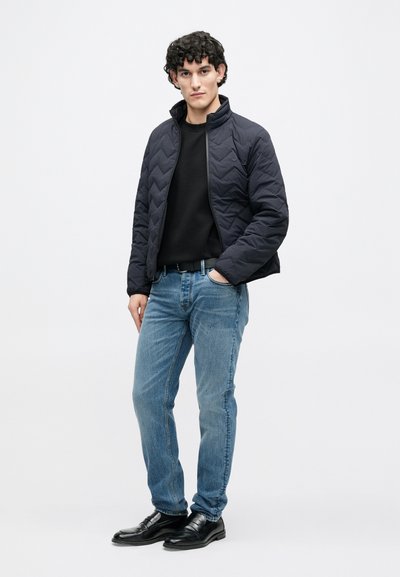 Tmavomodrá prešívaná bunda s vysokým golierom, pod ňou čierny sveter, svetlomodré rifle a čierne kožené topánky. Model stojí na neutrálnom pozadí.