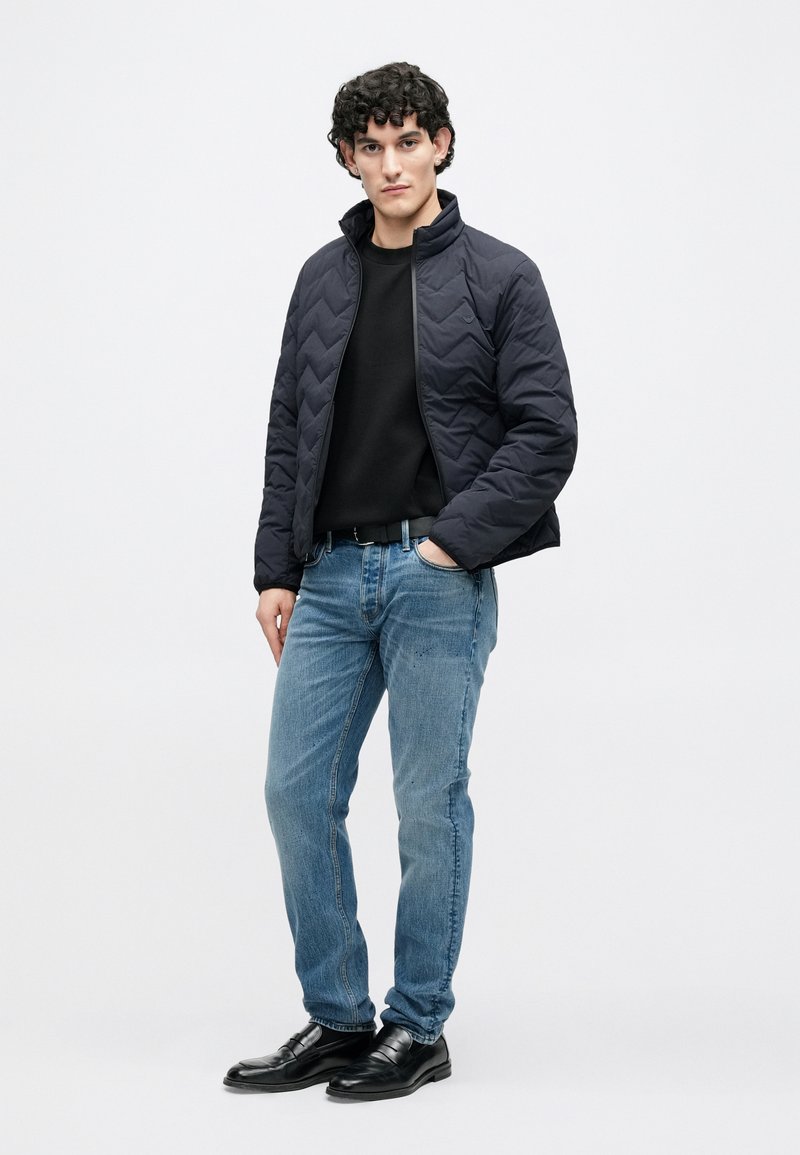 Veste matelassée marine avec col haut, pull noir en dessous, jean bleu clair et chaussures en cuir noir. Le mannequin est posé sur un fond neutre.