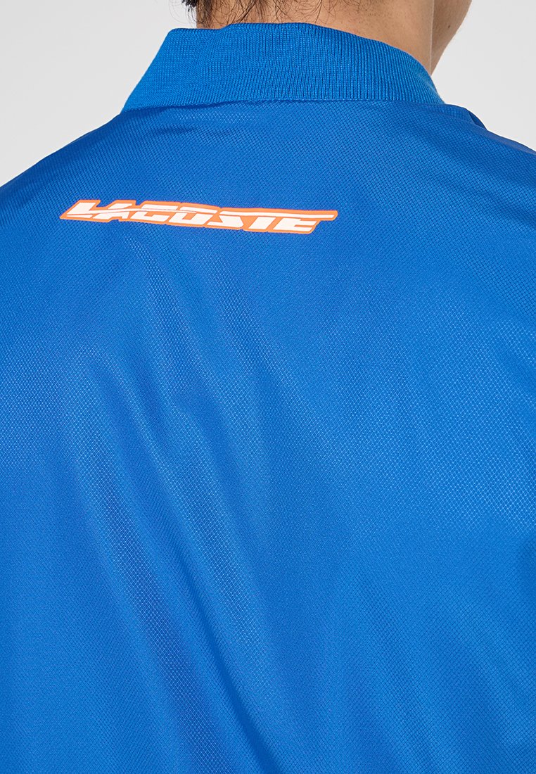 Polo azul hecho de tela texturizada, con cuello y logo "LACOSTE" en naranja en la parte posterior.