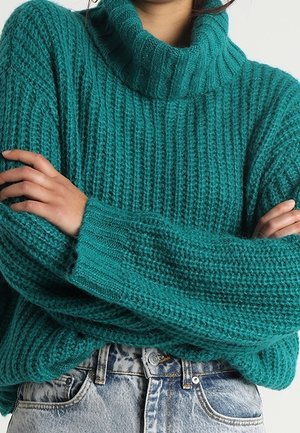 Person iført en kraftig mørk turkis rullekravetrøje med armene over kors, kombineret med lyseblå jeans med høj talje. Overkroppen vises.