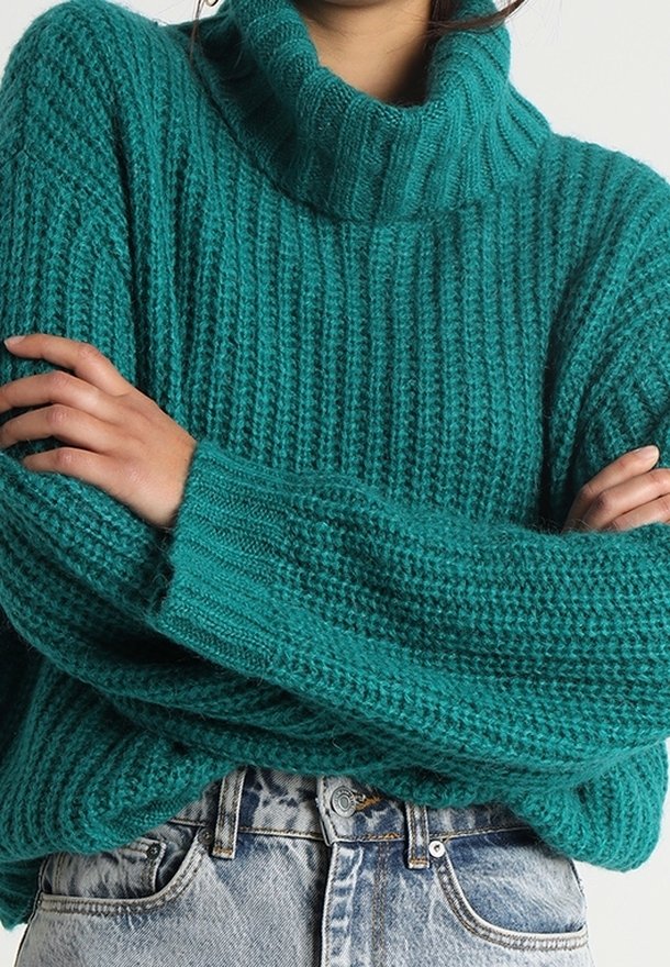 Personne portant un gros pull à col roulé bleu canard avec les bras croisés, assorti à un jean taille haute bleu clair. Le haut du corps est visible.