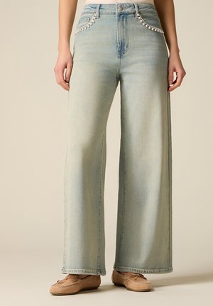 Jeans a gamba larga con lavaggio chiaro e bordatura di perline sulle tasche frontali, indossati con mocassini beige su una persona in piedi davanti a uno sfondo semplice.