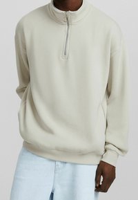 Uomo che indossa una felpa beige con zip a un quarto e anello, abbinata a jeans in denim azzurro, con la mano rilassata lungo il fianco, su uno sfondo neutro.