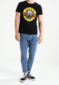 Zwart katoenen T-shirt met een kleurrijke "Guns N' Roses" grafiek met een schedel en rozen, gecombineerd met lichtblauwe jeans en zwarte sneakers.