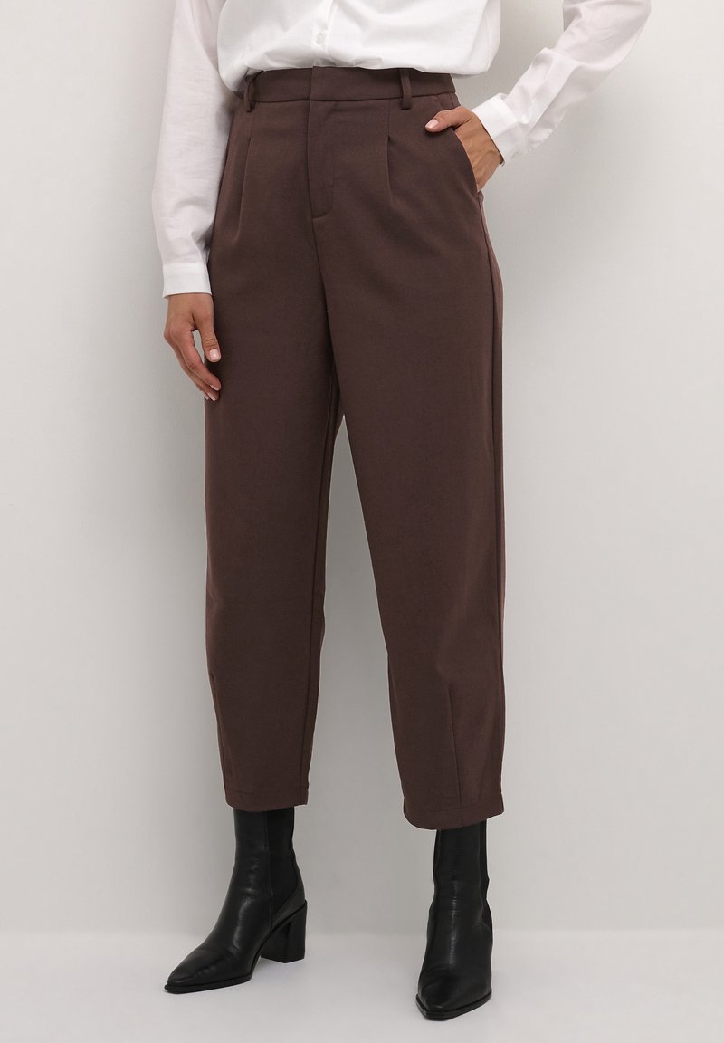 Femme portant un pantalon marron taille haute fuselé avec des plis à l'avant, une chemise blanche rentrée, et des bottines noires à talons, debout contre un mur blanc.