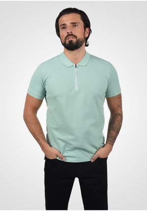 Camisa polo verde menta con detalle de cremallera, cuello tipo polo, mangas cortas y un diseño ajustado, combinada con pantalones negros. Tatuaje visible en el brazo.