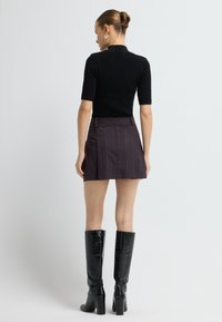 Kvinde med hår i en knold, iført sølvøreringe, en sort T-shirt med korte ærmer, en mørk lilla nederdel og sorte knæhøje støvler, stående med ryggen til.