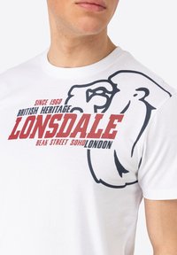 Balta medvilninė marškinėlių su juodu liūto kontūru ir raudonu užrašu "LONSDALE", su papildomais prekės ženklų detalėmis tamsiose raidėse.