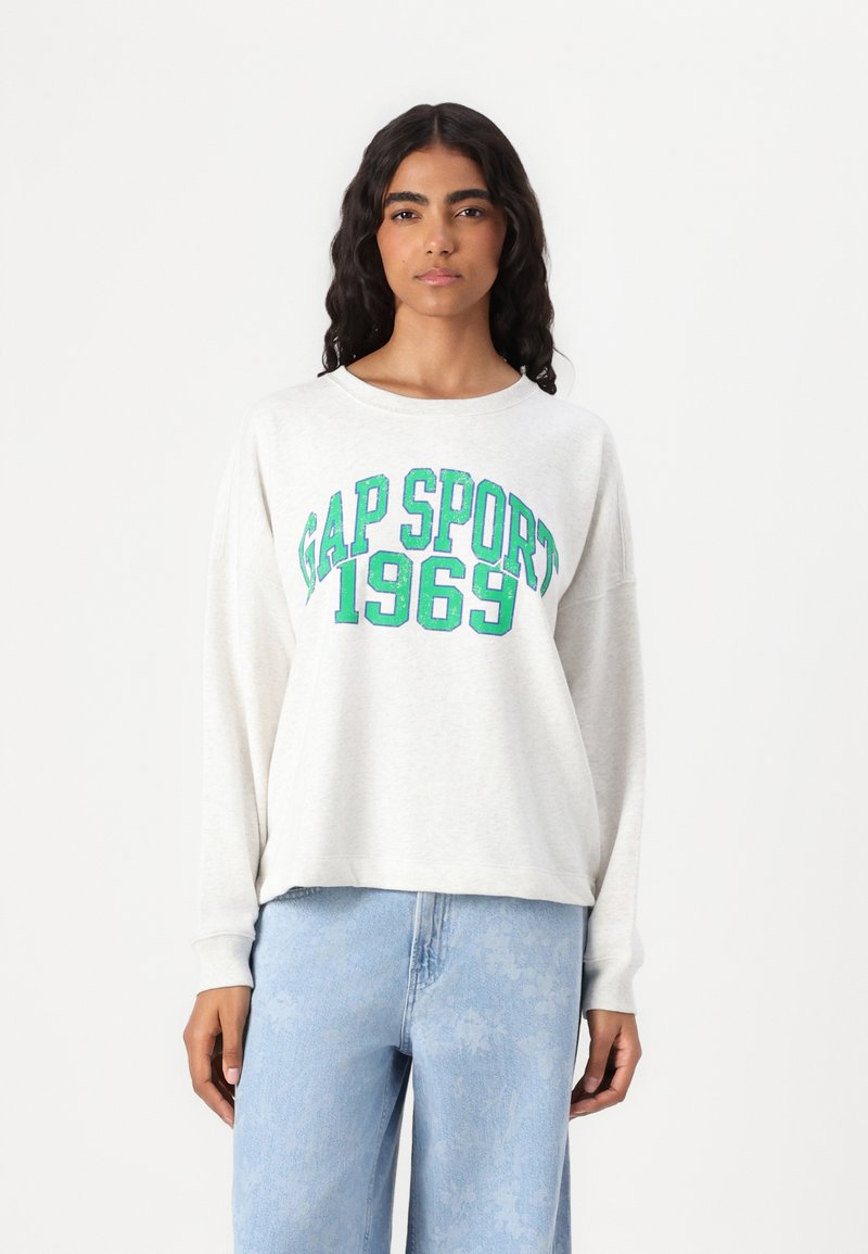 Felpa grigia chiara con la scritta "GAP SPORT 1969" stampata in verde, caratterizzata da una vestibilità comoda e maniche lunghe, indossata con jeans azzurri chiari.