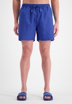 Shorts de bain bleus avec une taille élastique, un cordon de serrage et des poches latérales. Portés avec des sandales à glissière bleues assorties avec des accents blancs.