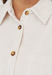 Cream ribbed corduroy shirt featuring a classic collar, two brown round buttons, and a chest pocket. Textured fabric with vertical lines.