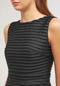 Robe noire avec un design texturé à rayures horizontales et de petites découpes. Style sans manches avec une silhouette ajustée.