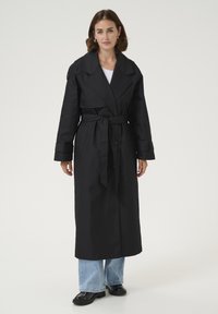 Cappotto lungo nero in un tessuto resistente, con un ampio colletto, vita con cintura e maniche lunghe. Indossato sopra jeans e scarpe nere lucide.