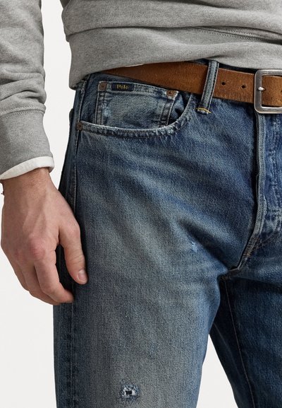Modri denim hlače z obrabljenim videzom, oblečene z rjavim usnjenim pasom, in sivo majico, ki je vidna blizu zapestja.