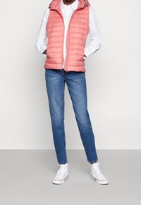 Rosa gepolsterte Weste mit Reißverschluss und Kapuze, getragen über einem weißen Shirt. Kombiniert mit blauen Jeans und weißen Sneakers. Glatte Textur, lässiges Design.