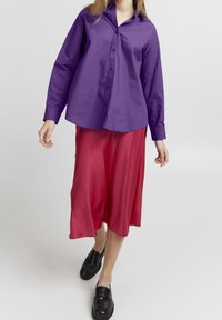 Lila Button-Up-Hemd mit langen Ärmeln, kombiniert mit einem fließenden roten Midirock und schwarzen Plateauschuhen, die kontrastierende Farben und Texturen präsentieren.