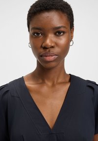 Mladá žena s krátkymi kučeravými vlasmi, nosiaca malé kruhové náušnice a tmavý top s výstrihom do V, pozerajúca sa priamo do kamery.