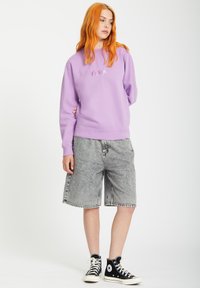 Liliowa bluza z ribbowymi mankietami, z subtelnym logo. W połączeniu z szarymi dżinsowymi szortami i czarnymi sneakersami za kostkę.