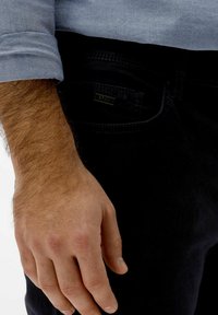 Jeans neri con una finitura testurizzata, dotati di un'etichetta metallica sulla tasca posteriore. Una mano si appoggia sul lato, mettendo in mostra il tono di pelle e i capelli naturali.