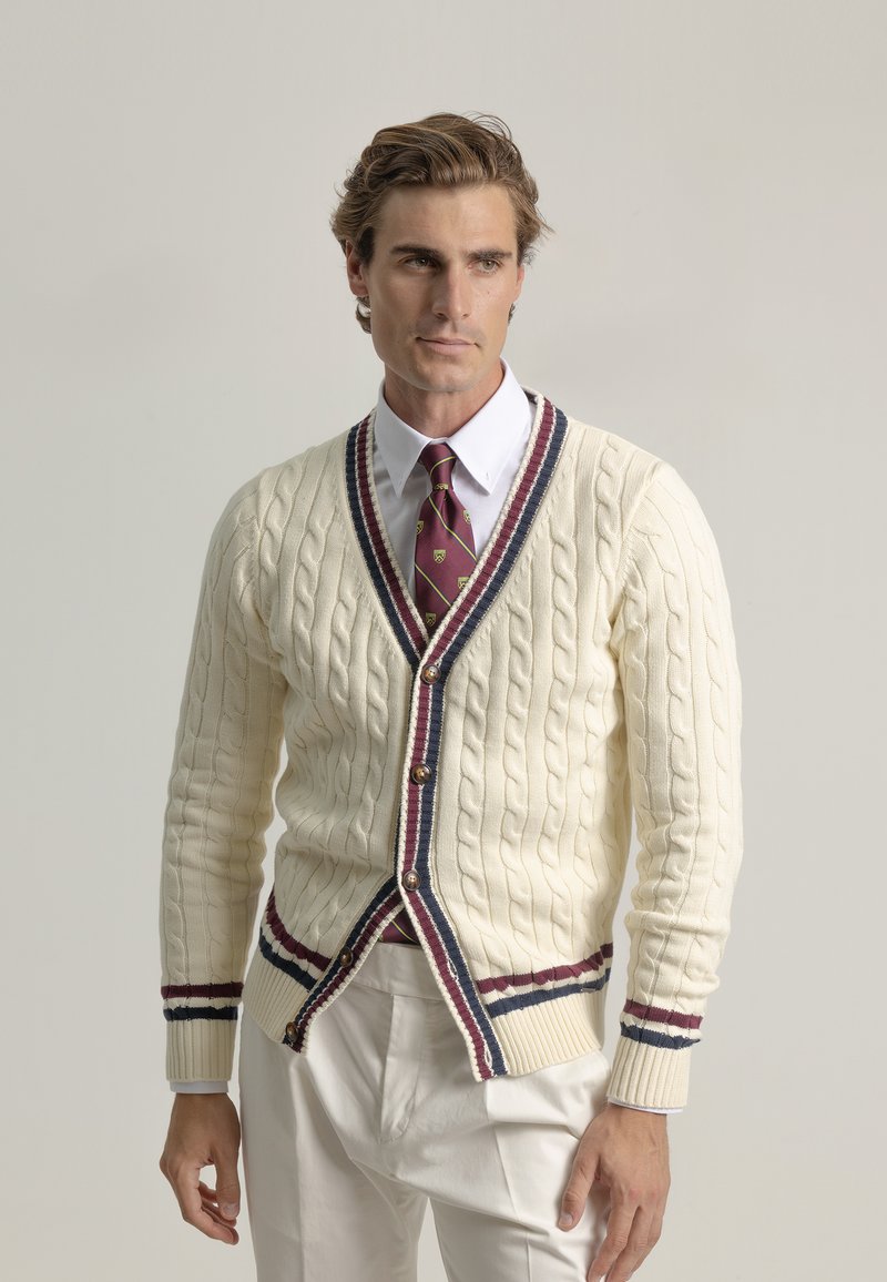 Suéter de punto crema con trenzado y escote en V profundo, con ribetes en contraste de color burdeos y marino, y botones de madera. Lleva puesto sobre una camisa blanca y una corbata.