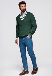 Maglione verde lavorato a maglia con scollo a V e accenti bianchi, abbinato a jeans azzurri chiari e scarpe marroni. Tessuto testurizzato con un motivo a rombi.