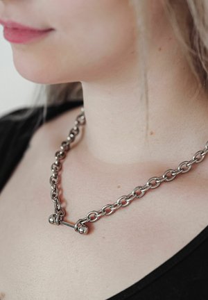 Nahaufnahme einer Person, die eine dicke silberne Kettenhalskette mit einem hantelartigen Verschluss trägt und ein schwarzes Oberteil anhat.