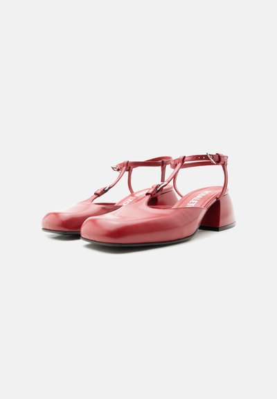 Κόκκινα δερμάτινα παπούτσια με λουράκι σε σχήμα Τ, χαμηλό τακούνι τύπου block, κλείσιμο με αγκράφα στον αστράγαλο, στρογγυλερή μύτη και επώνυμο πάτο σε λευκό φόντο.