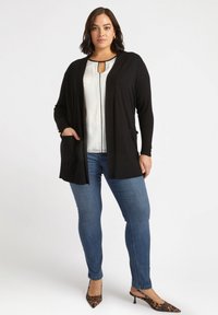 Schwarzer langer Cardigan mit offenem Vorderteil und Taschen, getragen über einem hellen Oberteil mit Schlüsselloch-Ausschnitt, kombiniert mit blauen Jeans und Leopardenmuster-Absätzen.