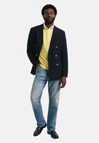 Marineblauwe gestreepte dubbelbreasted blazer over een gele sweater, gecombineerd met lichtblauwe spijkerbroek en zwarte nette schoenen.