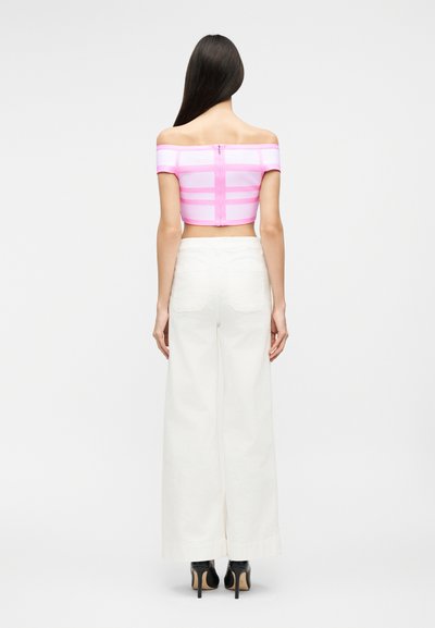 Γυναίκα με μακριά σκούρα μαλλιά, φορώντας ροζ τοπ με ακάλυπτους ώμους και λευκό, φαρδύ παντελόνι ψηλόμεσο σε φαρδιά γραμμή, με μαύρα ψηλοτάκουνα, στέκεται γυρισμένη με την πλάτη.