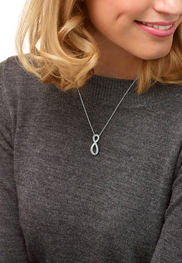 Sølv infinity vedhængsørering på en fin kæde, med et slankt, poleret design og glat tekstur, båret over en mørkegrå sweater.