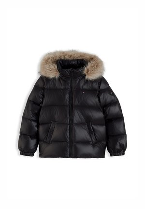 Ung person iført en sort dunjakke med pels hætte, hvid strikket sweater, mørke brede jeans og sorte sko, står med hænderne i lommerne.