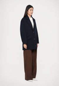 Meresinine mahukam villane jope, millel on suured kraed, ühe rea nööpkinnitused, küljetaskud ning mis on kombineeritud pruunide laiade pükstega ja valgete kingadega.