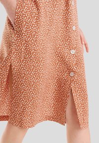 Vestido marrón con lunares blancos, que presenta una abertura lateral y botones en la parte delantera. Está hecho de una tela suave con una textura lisa.