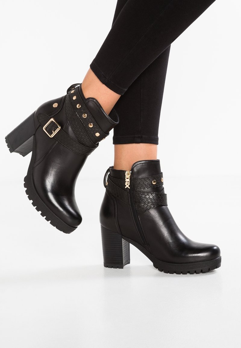 Bottines en cuir noir à talons blocs, fermeture éclair latérale, brides décoratives avec clous dorés et boucle, portées avec des leggings noirs.