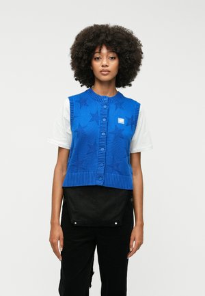 Sininen neulottu liivi, jossa on tähtikuvio, joustava helma ja napitus. Päällä valkoinen paita ja mustat housut.