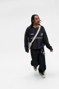 Mörkgrå stickad tröja med röda detaljer, löst sittande svarta jeans och grå sneakers. En beige axelväska och lager-på-lager halsband kompletterar outfiten.