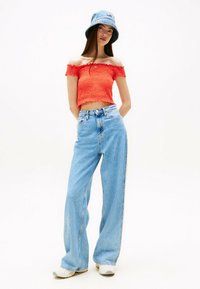 Crop top rosso a spalle scoperte con texture a coste, abbinato a jeans a gamba larga blu chiaro. La modella indossa un cappello a secchiello di denim blu.