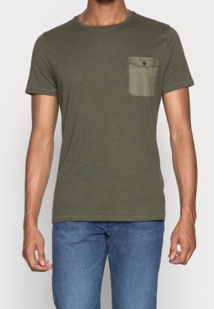 Homme portant un t-shirt vert olive à manches courtes avec une poche poitrine boutonnée et un jean bleu, debout devant un fond uni.