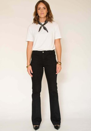 T-shirt blanc à manches courtes, jean flare noir taille haute, et talons aiguilles noirs. Foulard noir noué autour du cou.