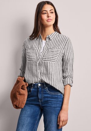 Młoda kobieta ubrana w szarą i białą koszulę zapinaną na guziki w pionowe paski, niebieskie dżinsy, trzymająca w lewej ręce złożoną brązową zamszową kurtkę.