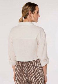 Kurz geschnittenes, hellbeiges Hemd aus strukturiertem Stoff, mit hochgerollten Ärmeln und einem Kragen. Darunter sind gemusterte, braune und weiße Hosen sichtbar.