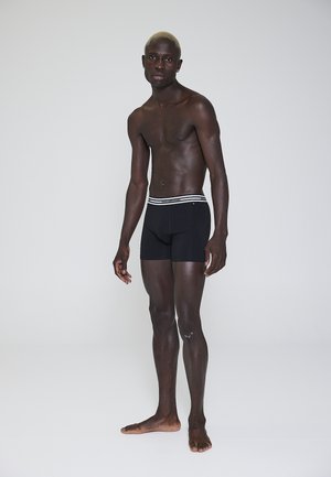Sorte boxer briefs med en visuelt kontrasterende hvidstribet elastikkant, lavet af et glat, strækbart stof. Modellen står barfodet.