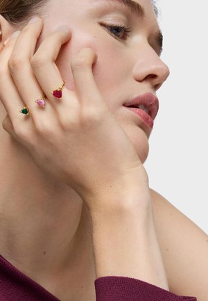 Main touchant le visage, portant trois bagues en or, chacune ornée d'une pierre en forme de cœur de couleur verte, rose et rouge, portant un haut violet.