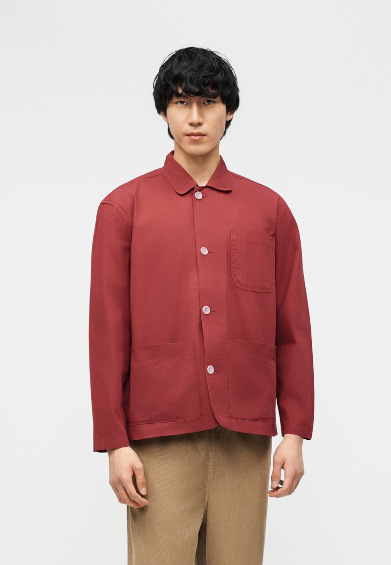 Άνδρας που φοράει κόκκινο μακρυμάνικο σακάκι με κουμπιά και τρεις μπροστινές τσέπες και μπεζ παντελόνι, στέκεται μπροστά σε απλό φωτεινό φόντο.