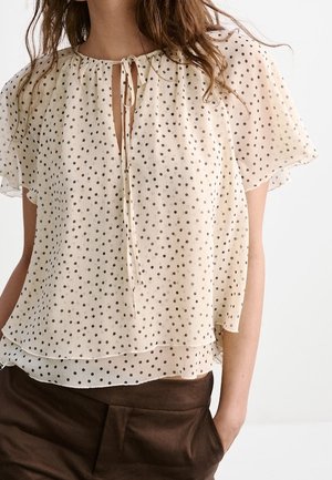 Femme portant un chemisier transparent beige clair à manches courtes avec des pois noirs et un col à nouer, associé à un pantalon marron foncé.